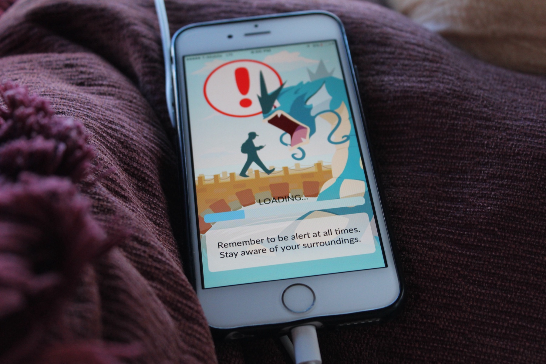 An iPhone resting on a blanket, showing the 2016 Pokémon GO loading screen with Gyarados and the reminder to stay aware of your surroundings.